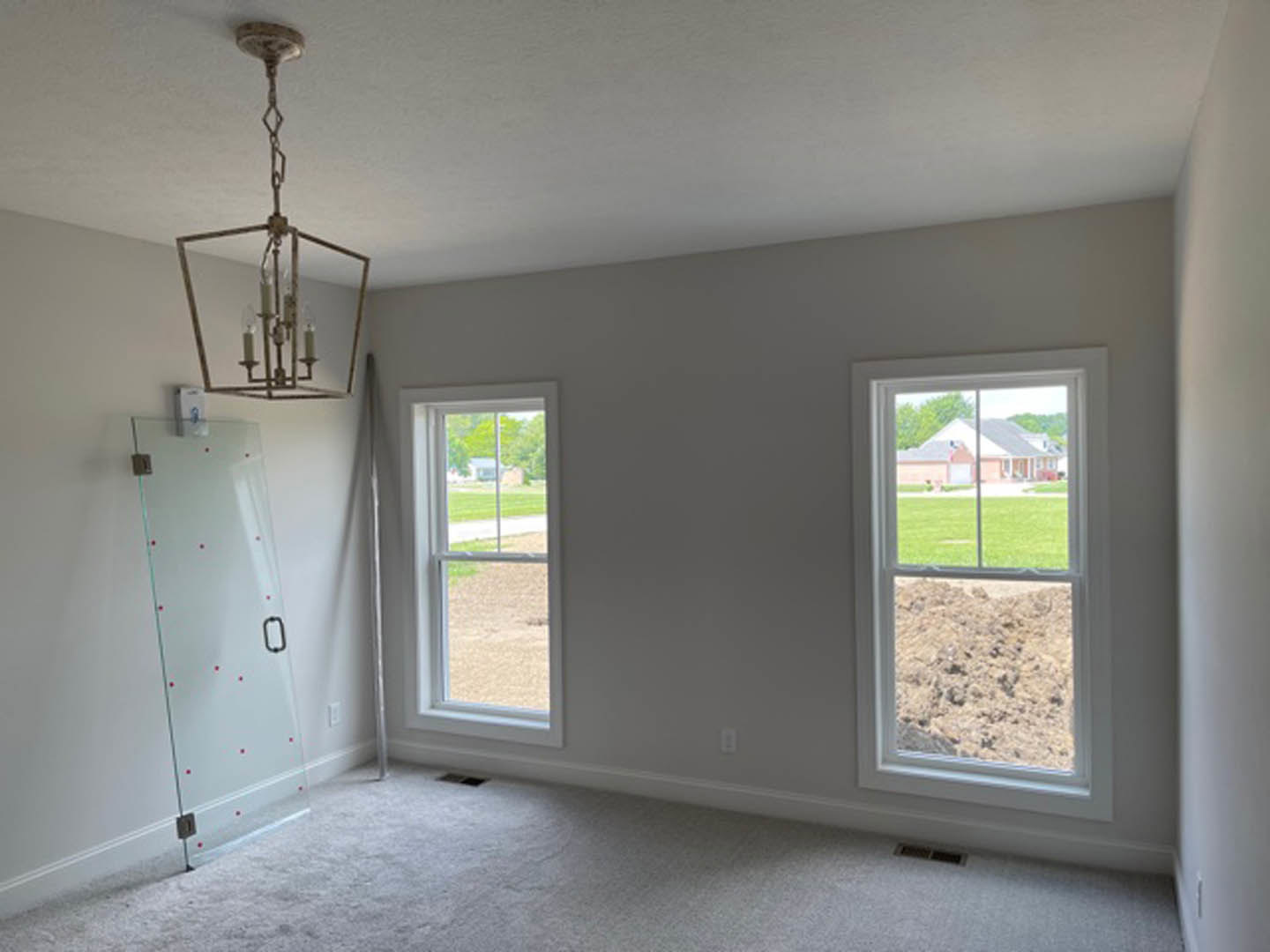 Bright room with large windows, glass door, white plaster walls, modern chandelier, beige carpet flooring, and outdoor views of grass, dirt field, and neighboring house