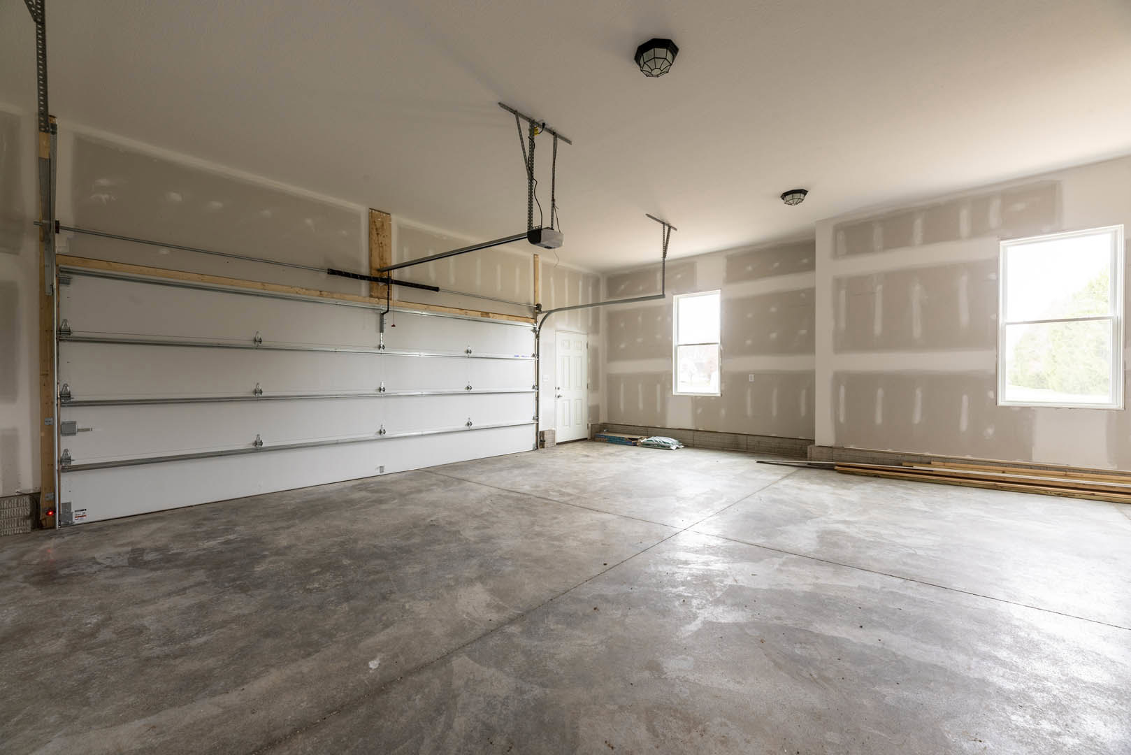 Empty room with white plaster walls and ceiling, concrete floor, white-framed window, white garage door with metal rails, and exposed metal pole with ceiling wires