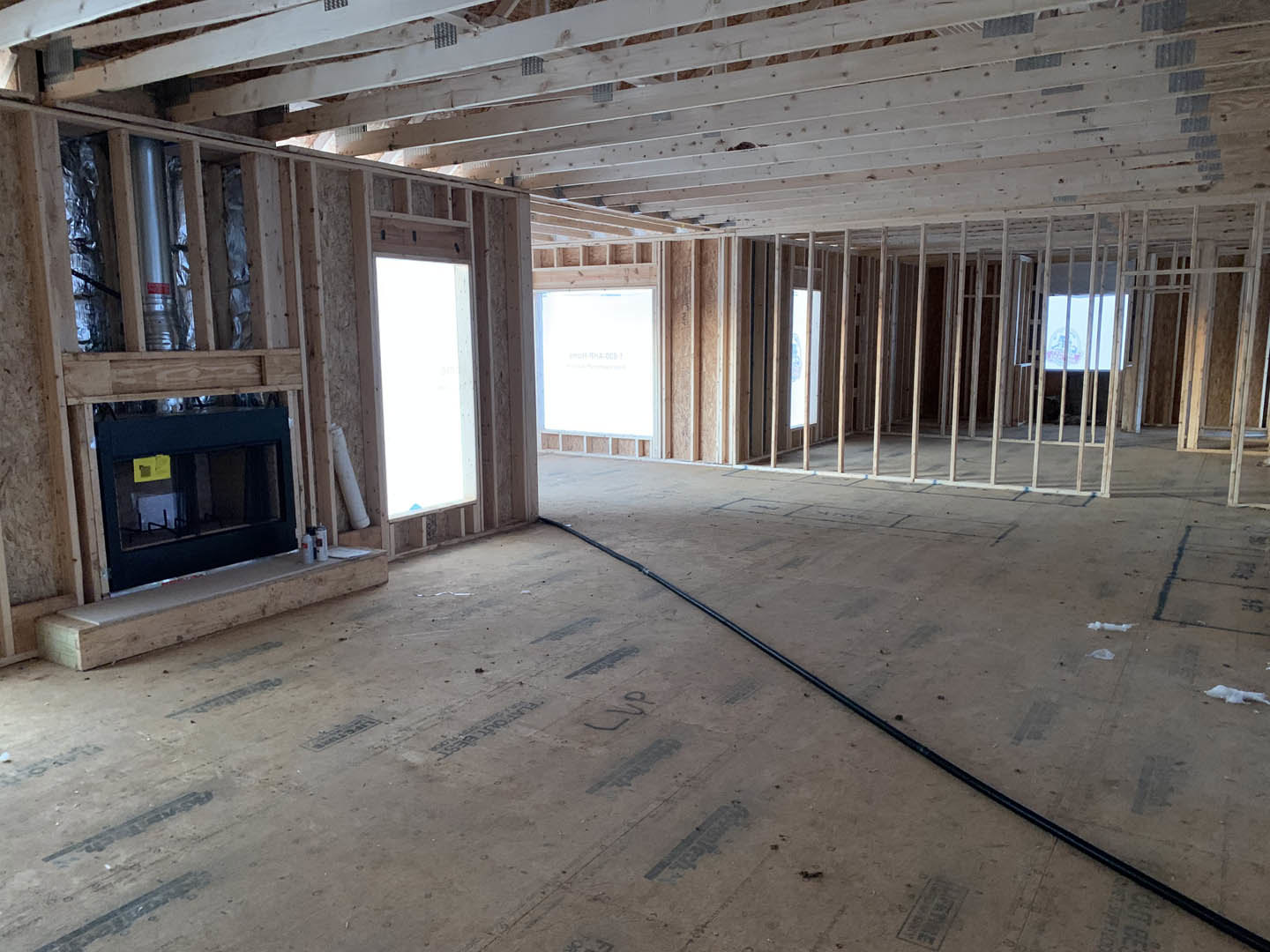 Living room with exposed wood ceiling beams, black fireplace with glass front, light wood floors, and large rectangular window.