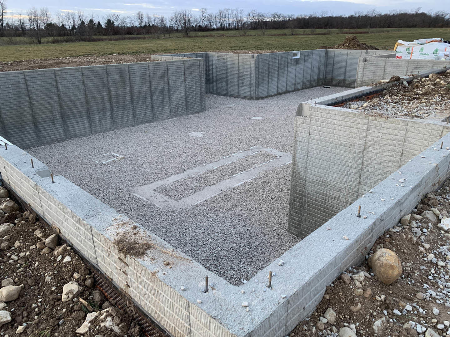 Concrete foundation surrounded by gravel, round rocks embedded in dirt, grey brick wall, sparse plants visible along outdoor ground.