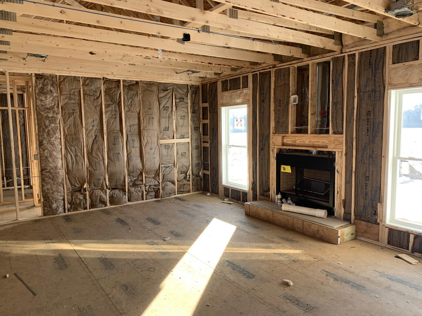 Living room with exposed wood ceiling beams, black fireplace, white-framed window overlooking a field, unfinished wall with insulation, and a countertop holding a roll of paper