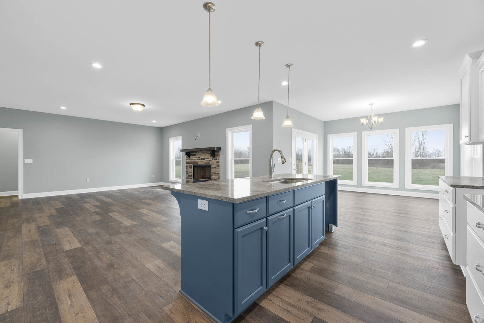 Open kitchen featuring a central fireplace with a wood mantel, marble-topped island, light cabinetry, and large window overlooking grassy field.