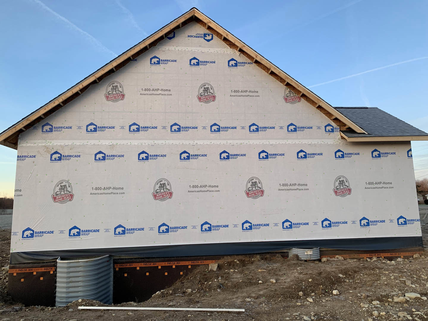 Framed house under construction with completed shingle roof, exposed plywood walls, and dormer window against blue sky