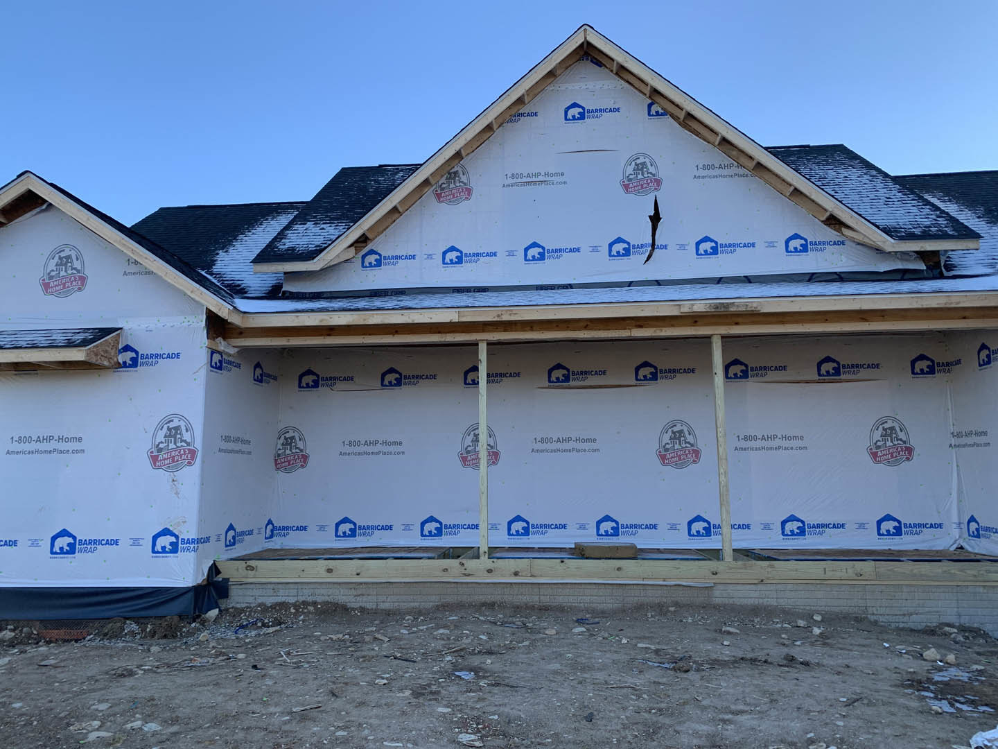 Partially built house with completed triangular roof, exposed framing, cracked exterior wall, dirt ground scattered with rocks and debris, white surface displaying a logo
