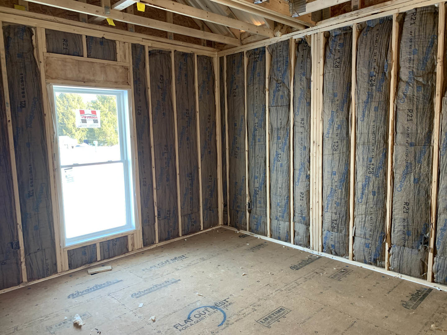Exposed wood ceiling beams above unfinished floor, large window framed by insulated walls, natural light entering construction space