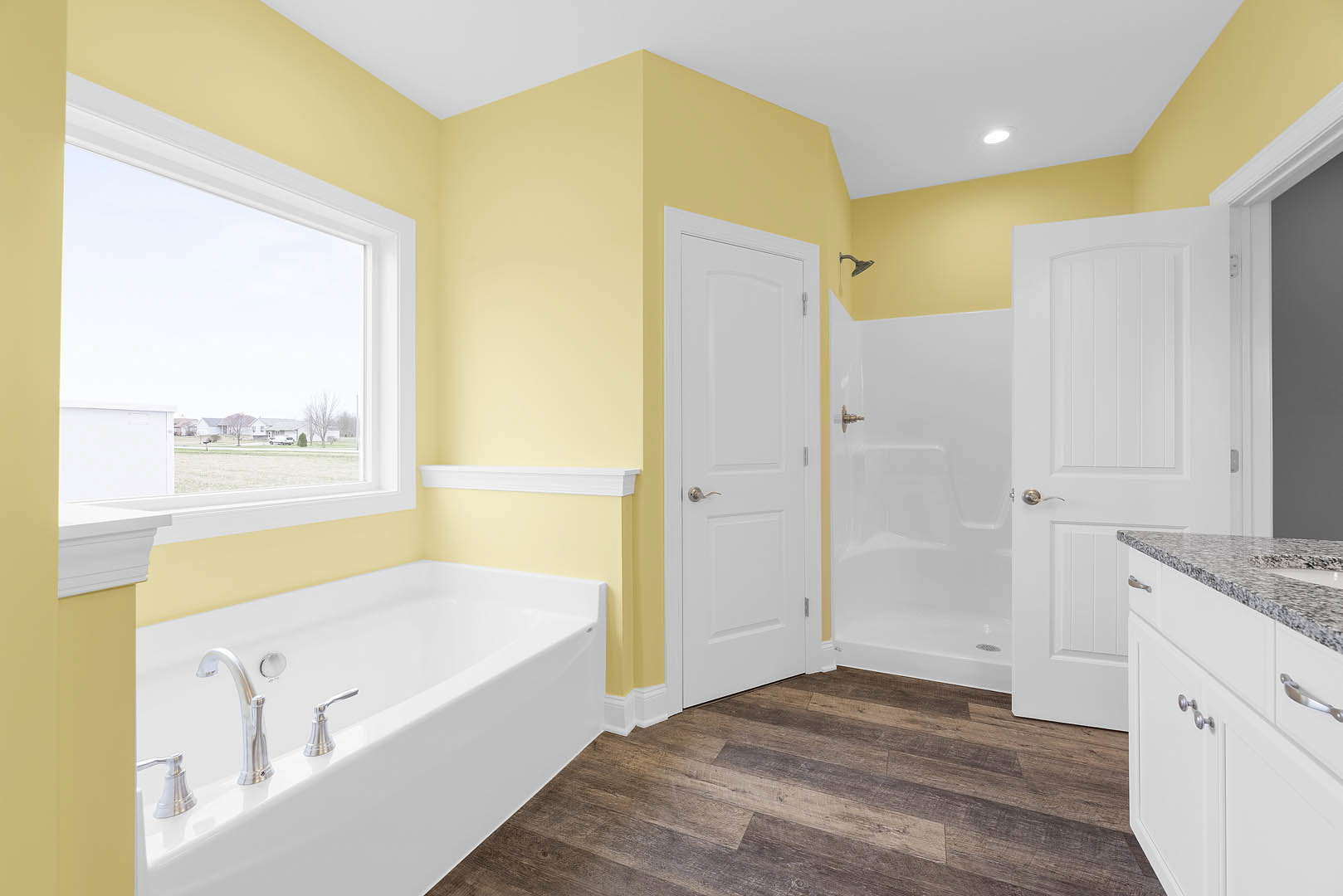 Bathtub with chrome faucets against a yellow wall, large window overlooking field and houses, wood flooring, white door with silver handle, kitchen counter visible in foreground