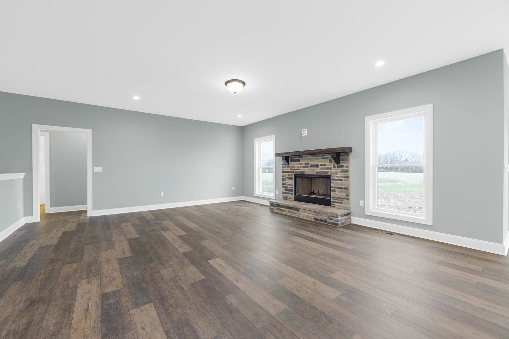 Living room with hardwood flooring, white plaster walls, glass-door fireplace topped by a wood mantel, ceiling light fixture, and window overlooking a grassy field
