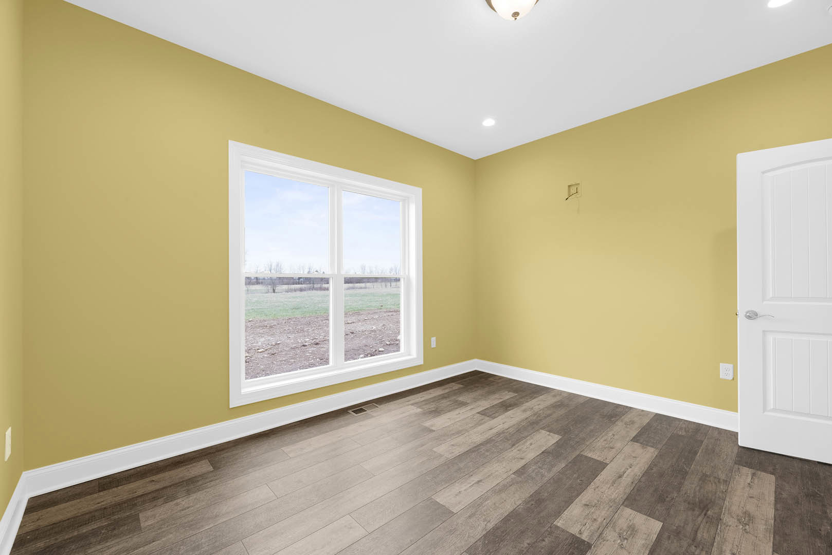 Sunlit room featuring wide plank wood flooring, white plaster walls, a white door with molding, and a large window overlooking a dirt field.