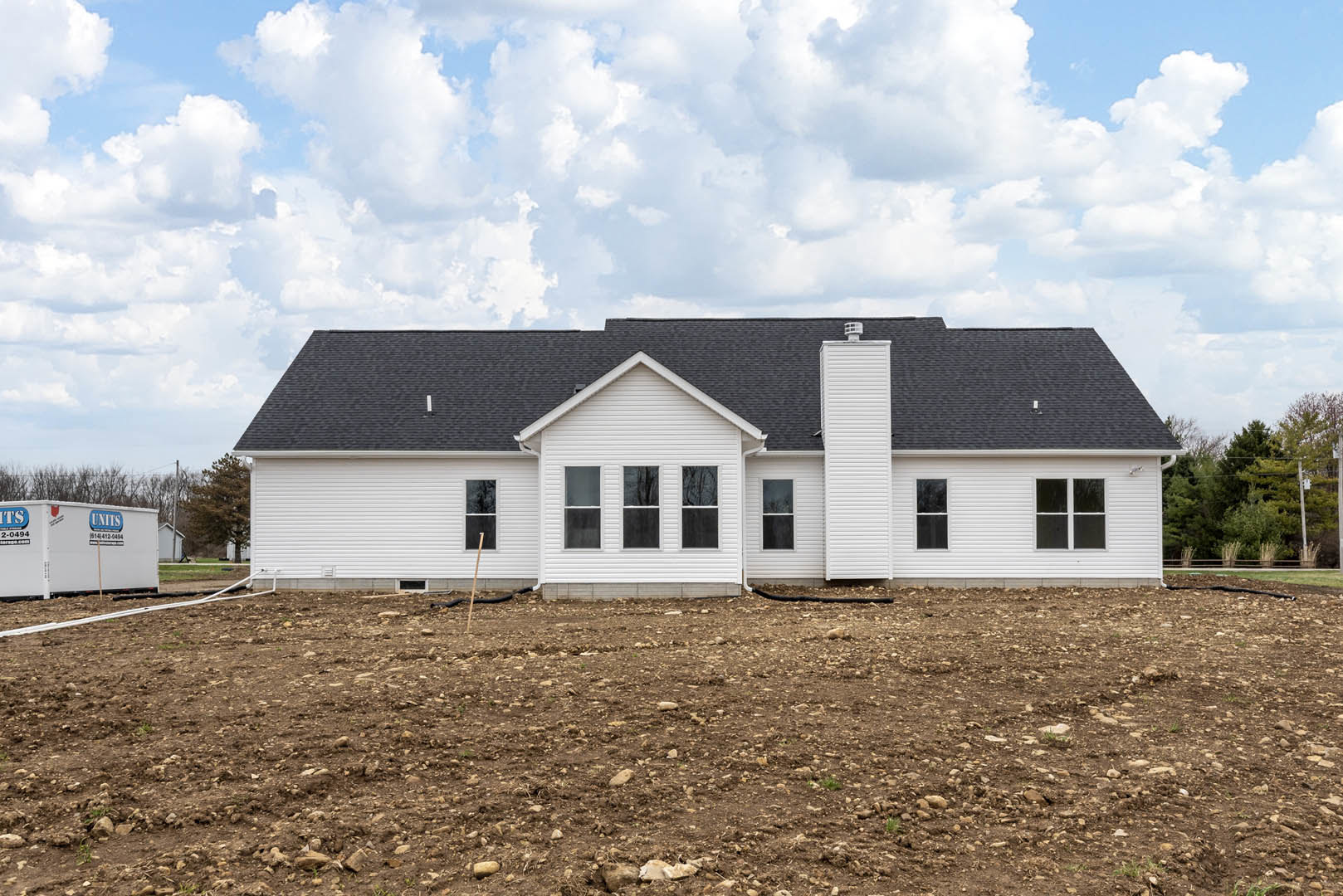 White house with black roof set beside a dirt field, white container with blue and white logos, black pipes near a white wall, cloudy sky overhead.