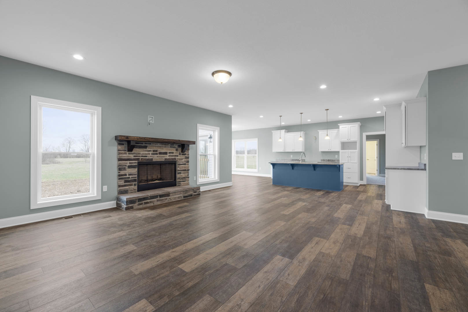 Spacious living room with hardwood floors, stone fireplace topped by a wood mantel, open kitchen area, large window overlooking a field, white door with black handles, modern