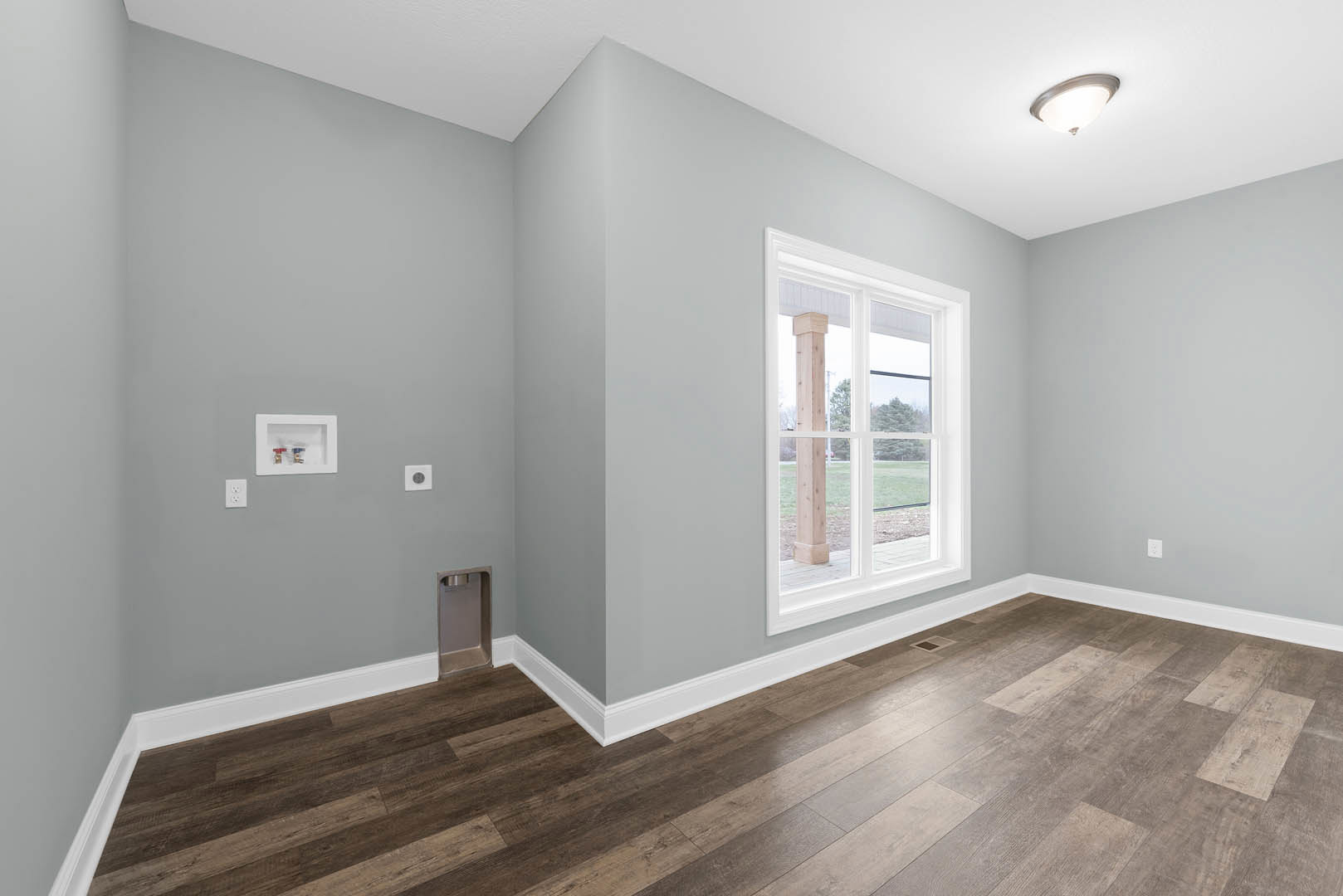 Sunlit room featuring wide wood flooring, white wall trim, large window with a central pillar, recessed white utility box with red and blue valves, rectangular metal-framed