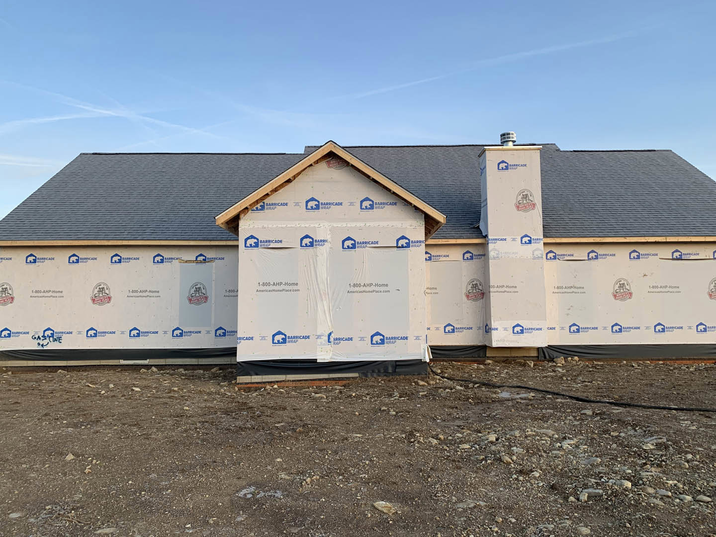 Two-story house under construction with blue and white insulation panels, exposed roof framing, dirt and rocks covering the ground, white pillars featuring blue and white logos.