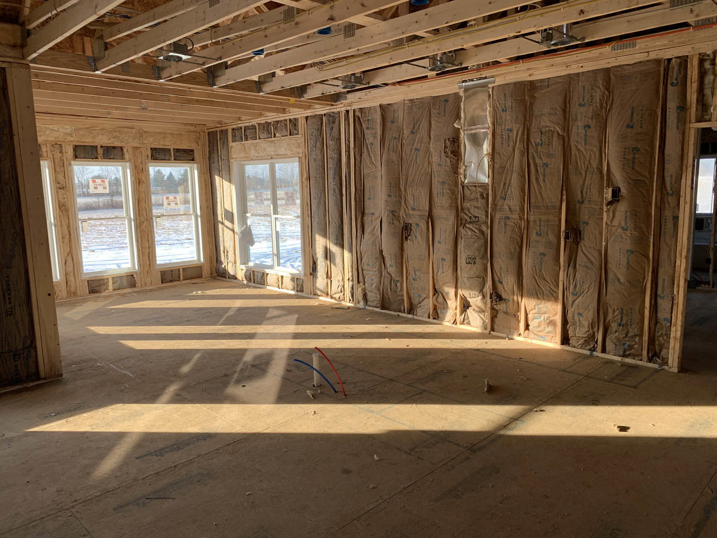 Room with exposed wood beams, unfinished walls covered in construction paper, large windows, visible pipes and insulation, white pole with blue and red tubes, close-up views of