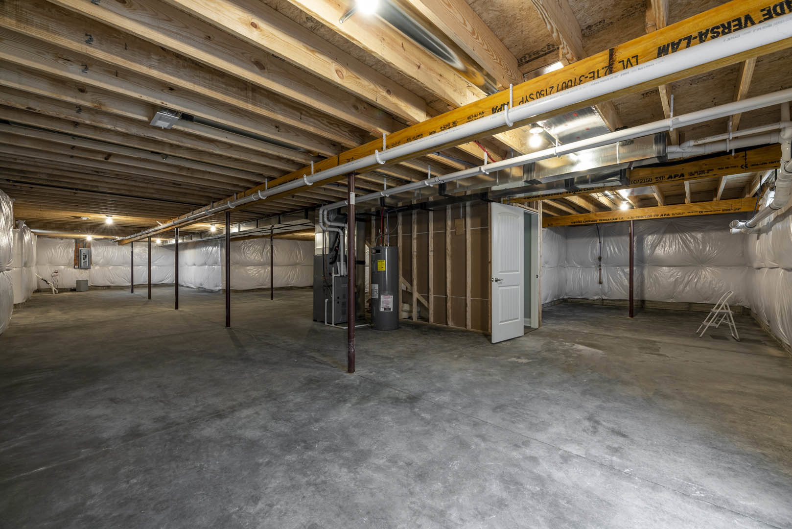 Concrete-floored room with exposed ceiling pipe, structural column, white door with silver handle, and white ladder on wood floor