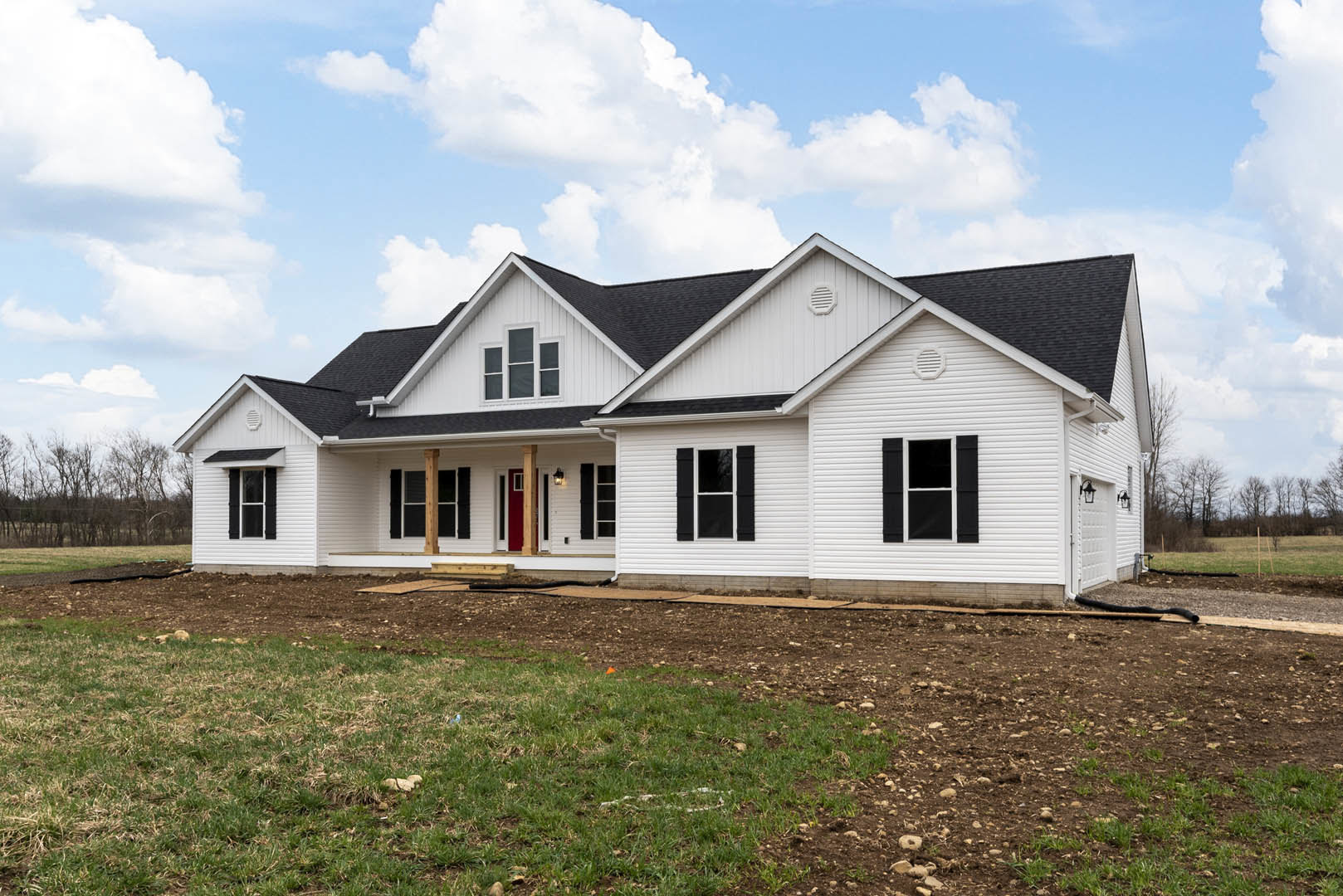 White siding house with black shuttered windows, red front door, green lawn, dirt patch, and blue sky with clouds