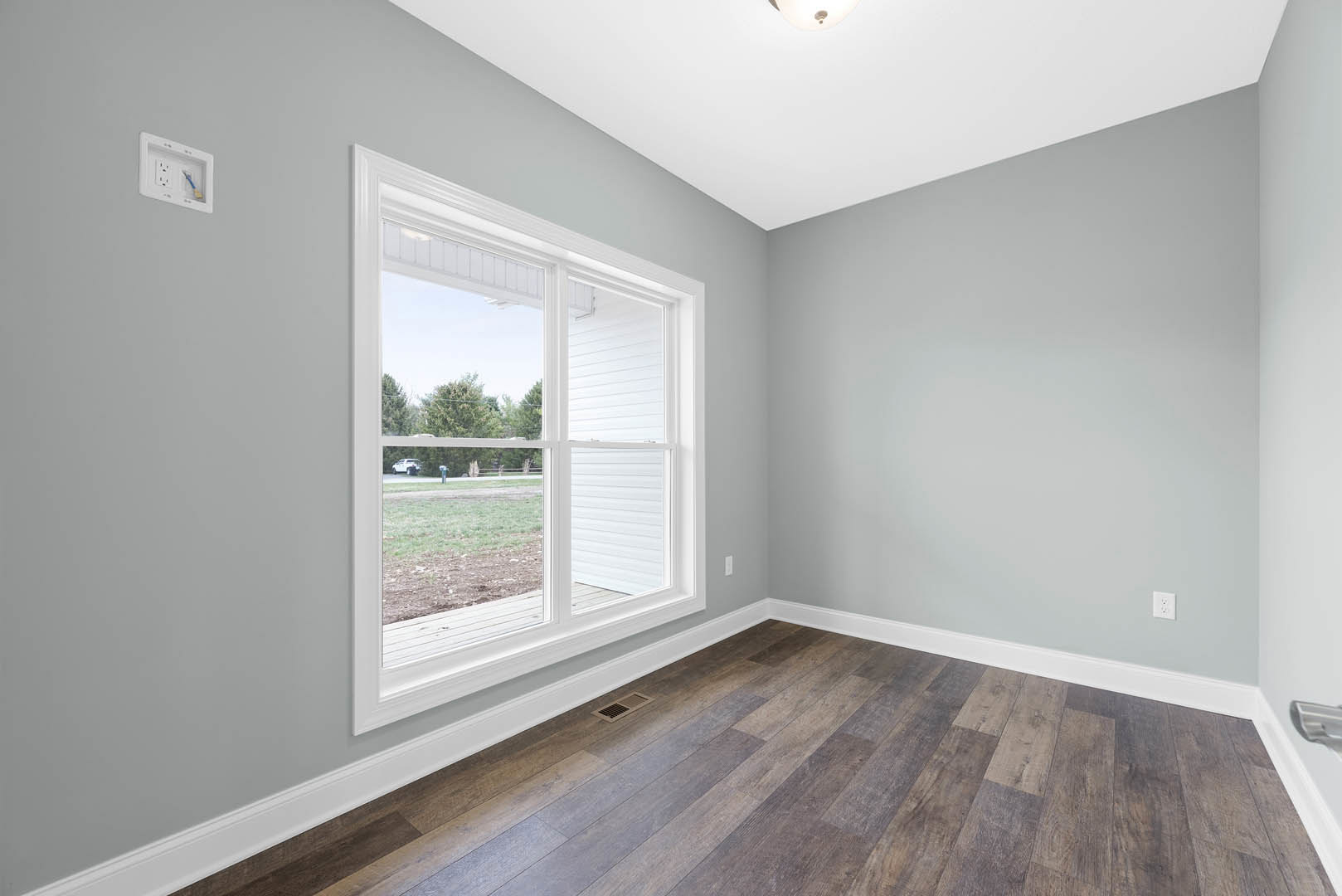 Sunlit room featuring a large window overlooking a grassy yard, white plaster walls with a recessed wall outlet, hardwood flooring with a vent in the corner