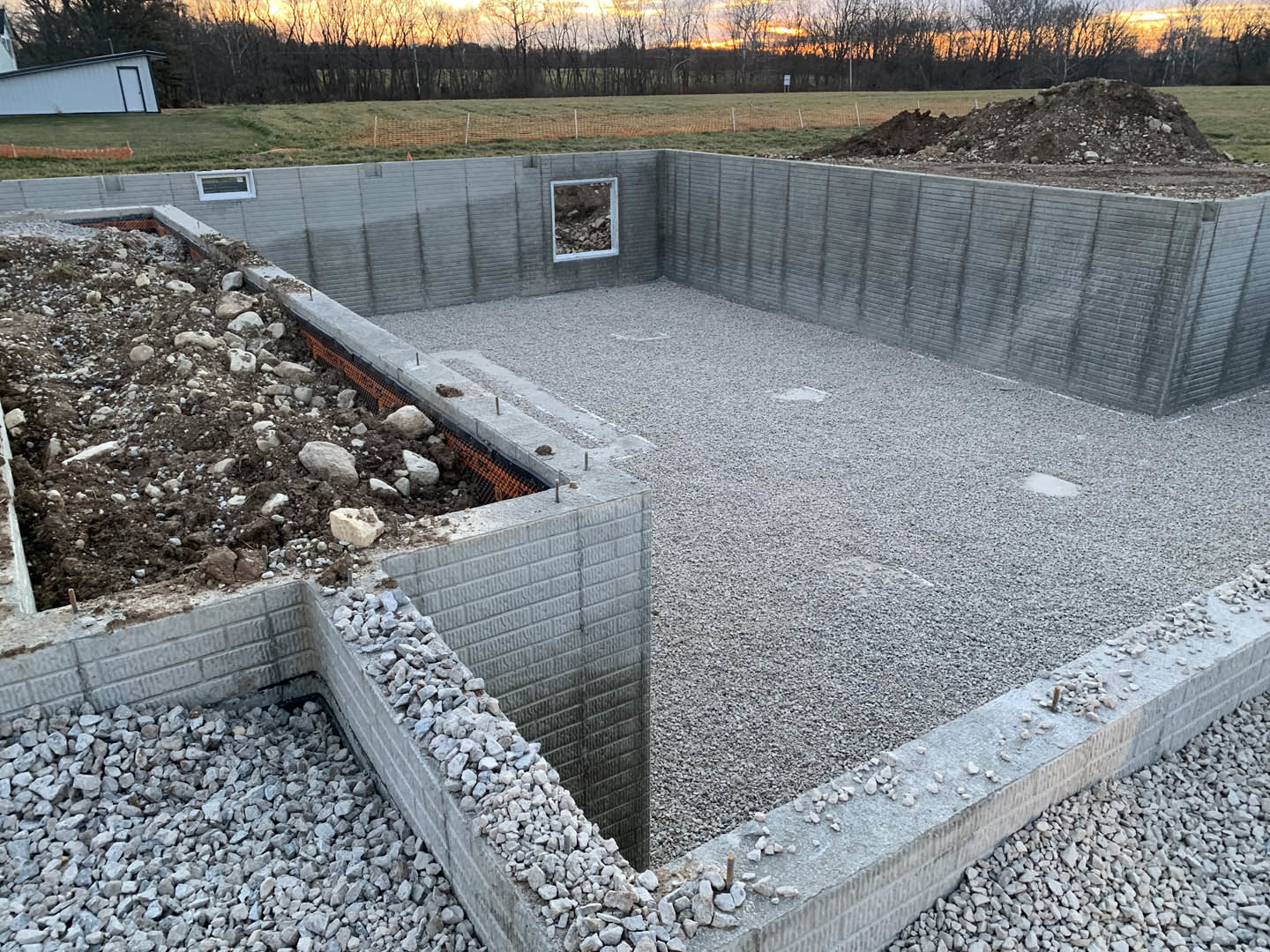 Concrete foundation surrounded by gravel, large rock on ground, pile of dirt and rocks atop concrete wall, white trailer with blue sign, sunset sky behind tree