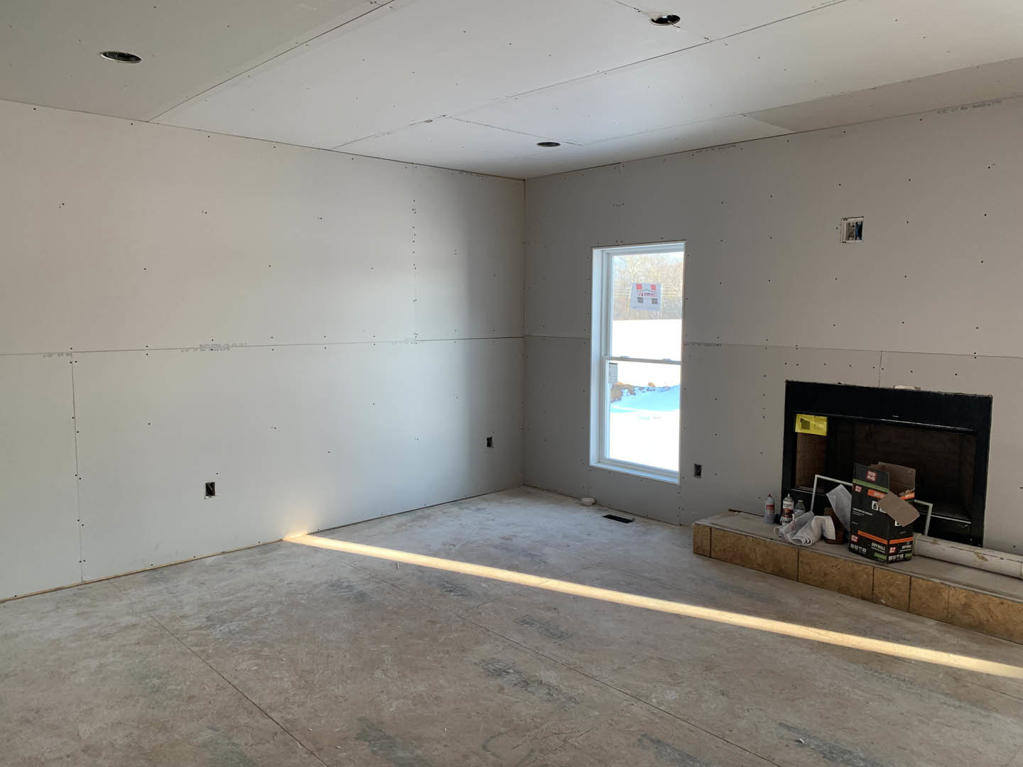 Living room with plaster walls, concrete floor marked by a white line, fireplace with black surround, large window framing snowy landscape, white ceiling with recessed holes