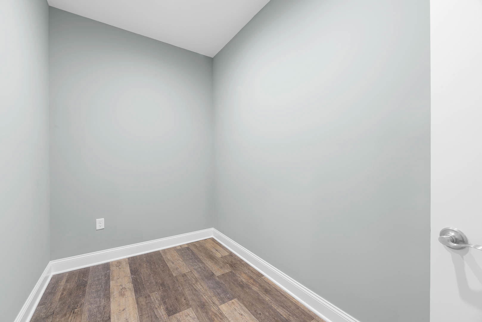 Corner of a room with wood flooring, white baseboards, white walls, and a white ceiling with a grey surface; close-up of a door handle visible.