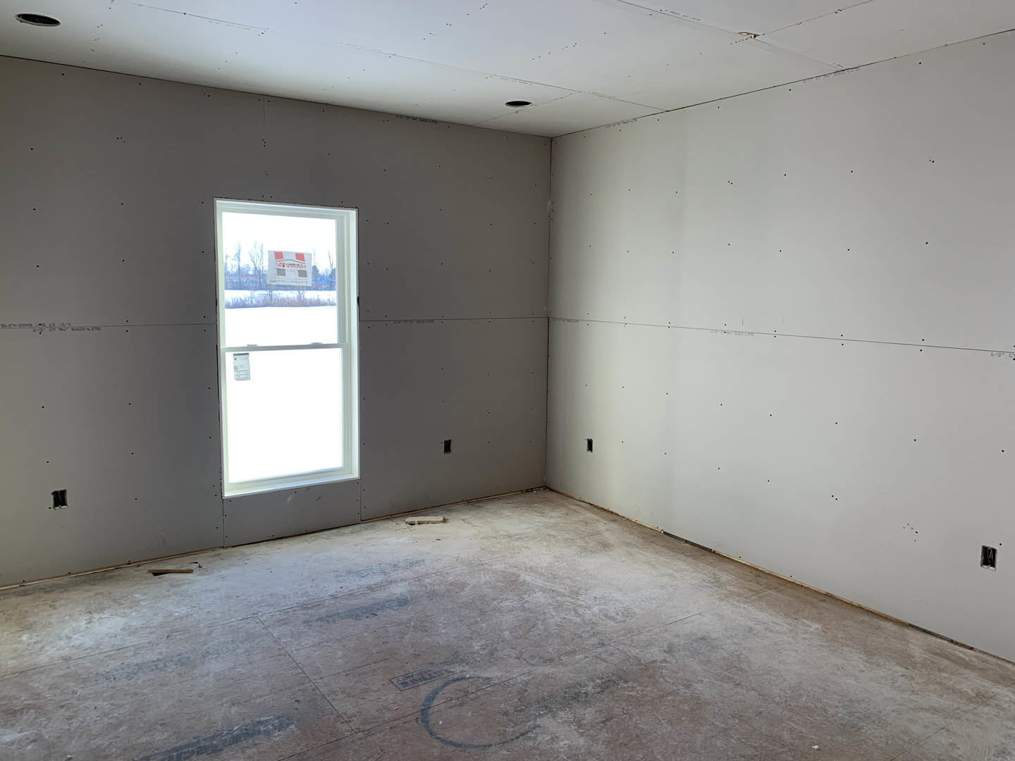 Concrete floor with scattered debris, large window featuring a sign, white ceiling with recessed holes, plaster walls, natural daylight illuminating the unfinished room