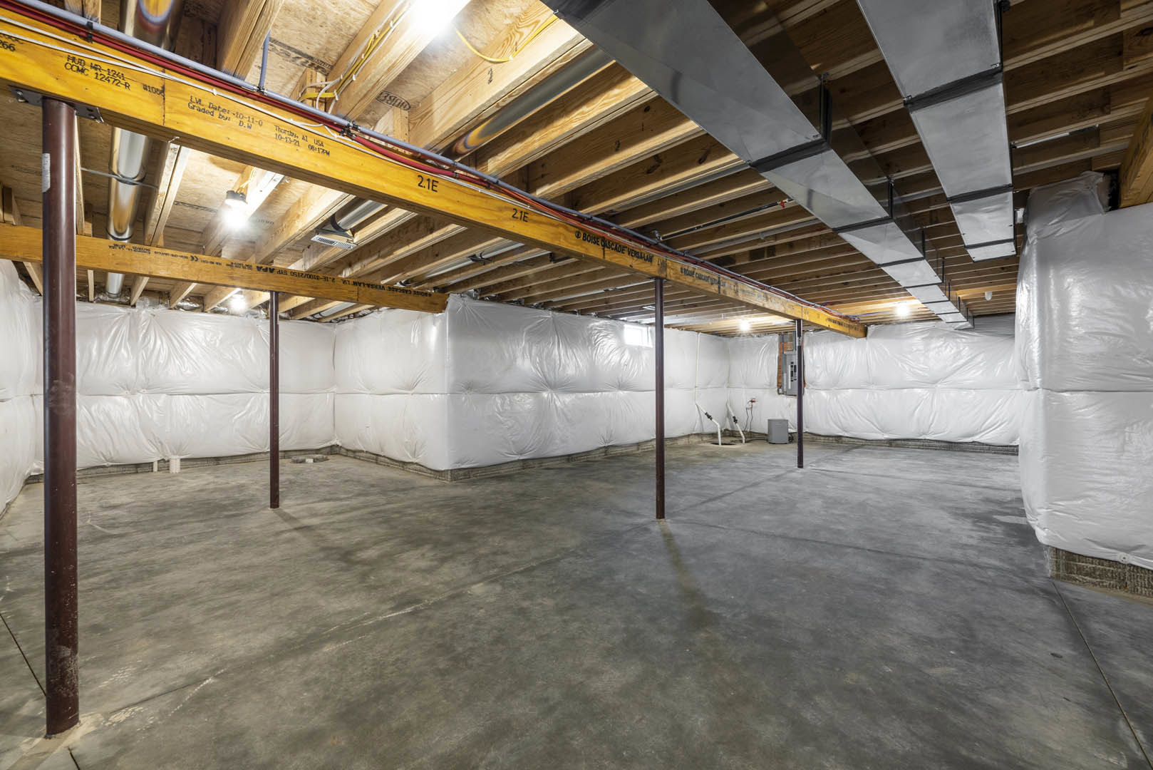 Exposed wooden ceiling beams with visible wiring and white pipes, concrete floor, white walls, and brown cylindrical object in an empty room