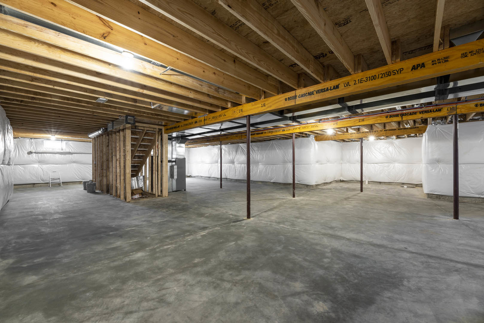 Open room with exposed wooden ceiling beams, concrete floor, steel support poles, and a white staircase against a light wall