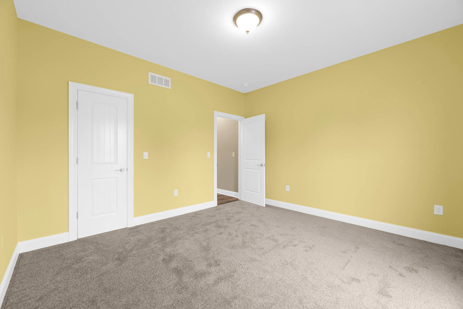 Yellow walls with white trim, white door featuring a silver handle, ceiling-mounted light fixture, carpeted floor, wall vent, and adjacent light switch.