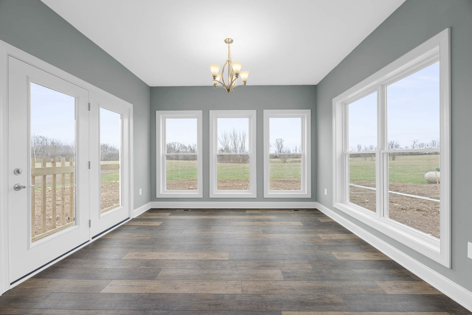 Spacious room with wood flooring, white walls, large windows overlooking grass and trees, glass doors, and an ornate chandelier hanging from the ceiling