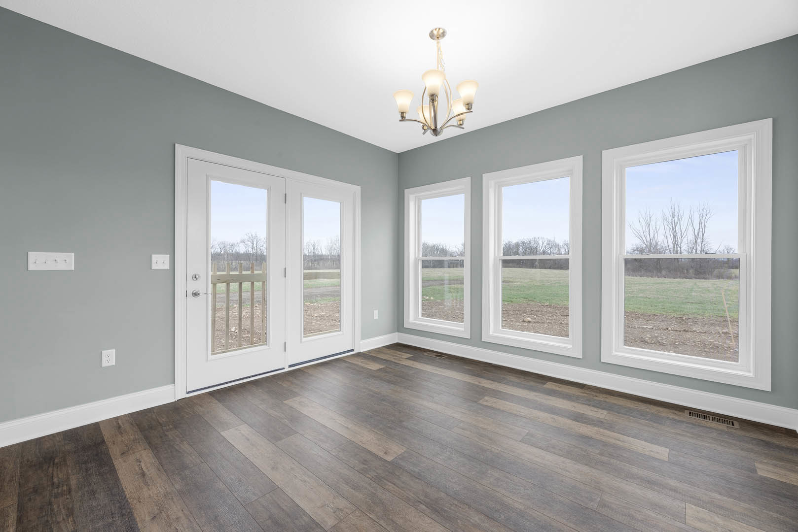 Wood flooring with white double doors featuring glass panels, large windows overlooking a field and trees, white walls, and a modern chandelier.