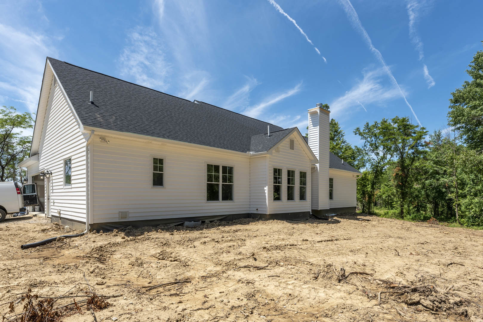 White house with black roof under construction, surrounded by dirt and trees, featuring white-framed windows and a white truck parked nearby.