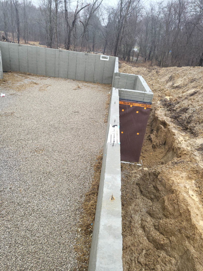 Concrete wall with circular hole in the ground, gravel and grass alongside, trees and forest visible in background