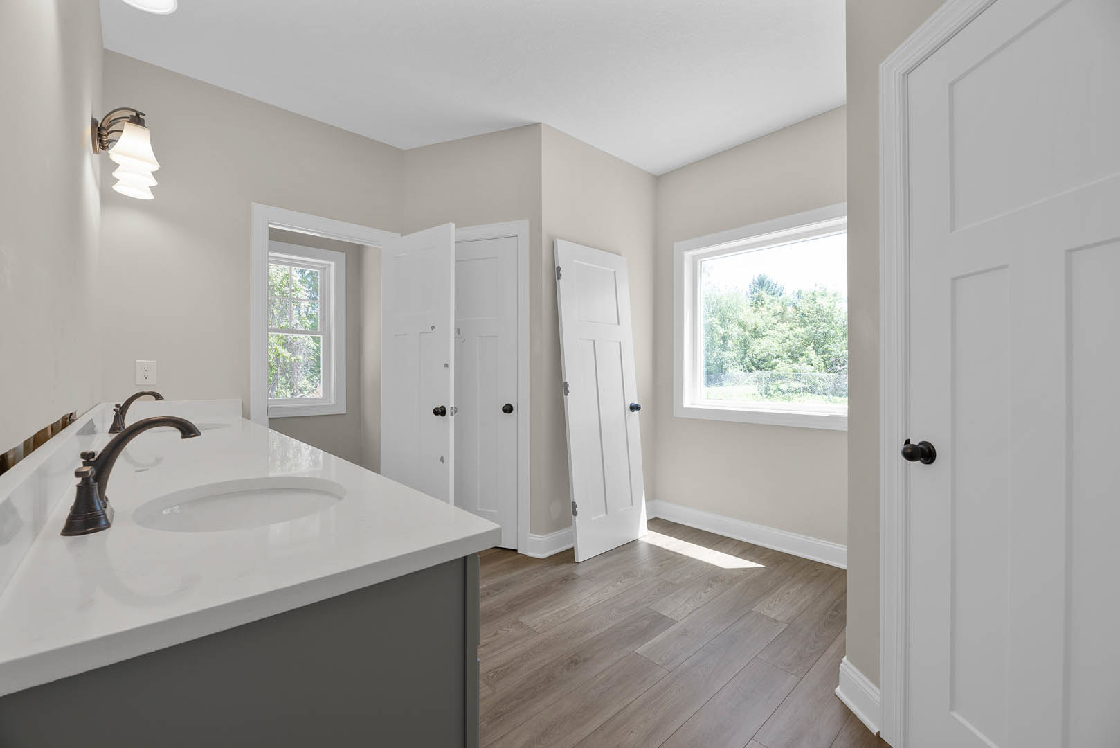 Bathroom with wood flooring, white sink and chrome faucet, window overlooking trees, white door with blue knob, wall-mounted light fixture with white shade