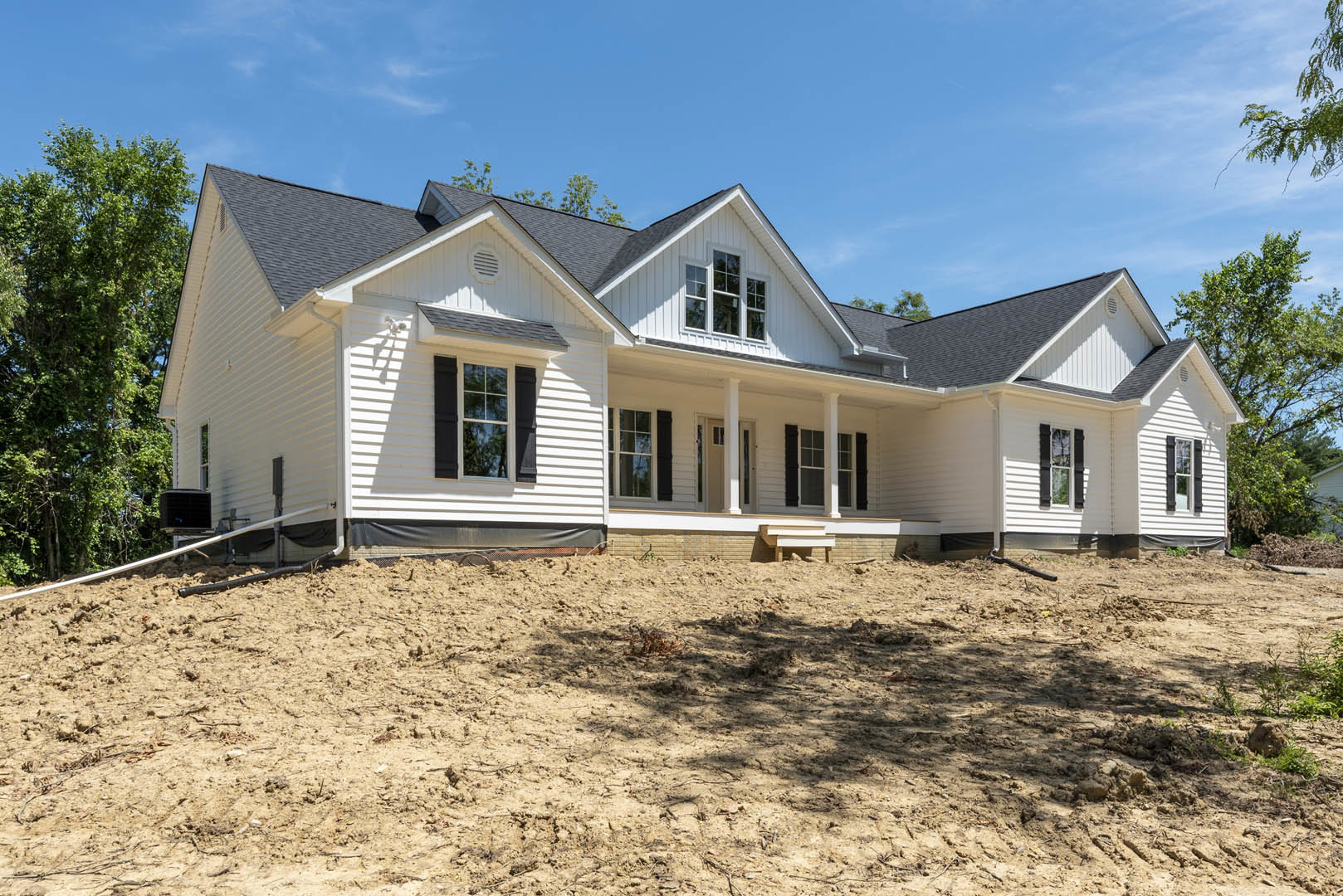 Two-story house with white siding, black shutters, and covered porch, set beside a dirt hill; mature tree with green leaves in foreground, Robert Frost Farm visible in background.
