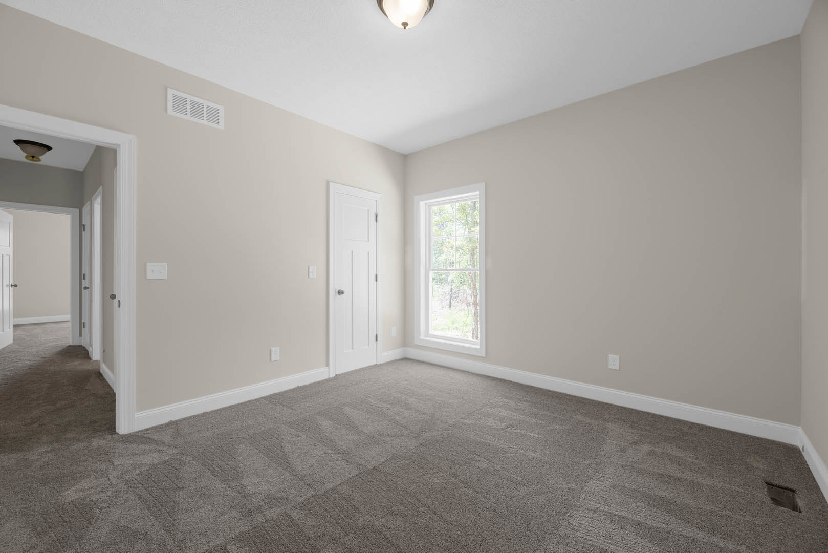 White carpeted room with a white paneled door featuring silver knobs, large window overlooking trees, ceiling-mounted light fixture, and smooth plaster walls with decorative
