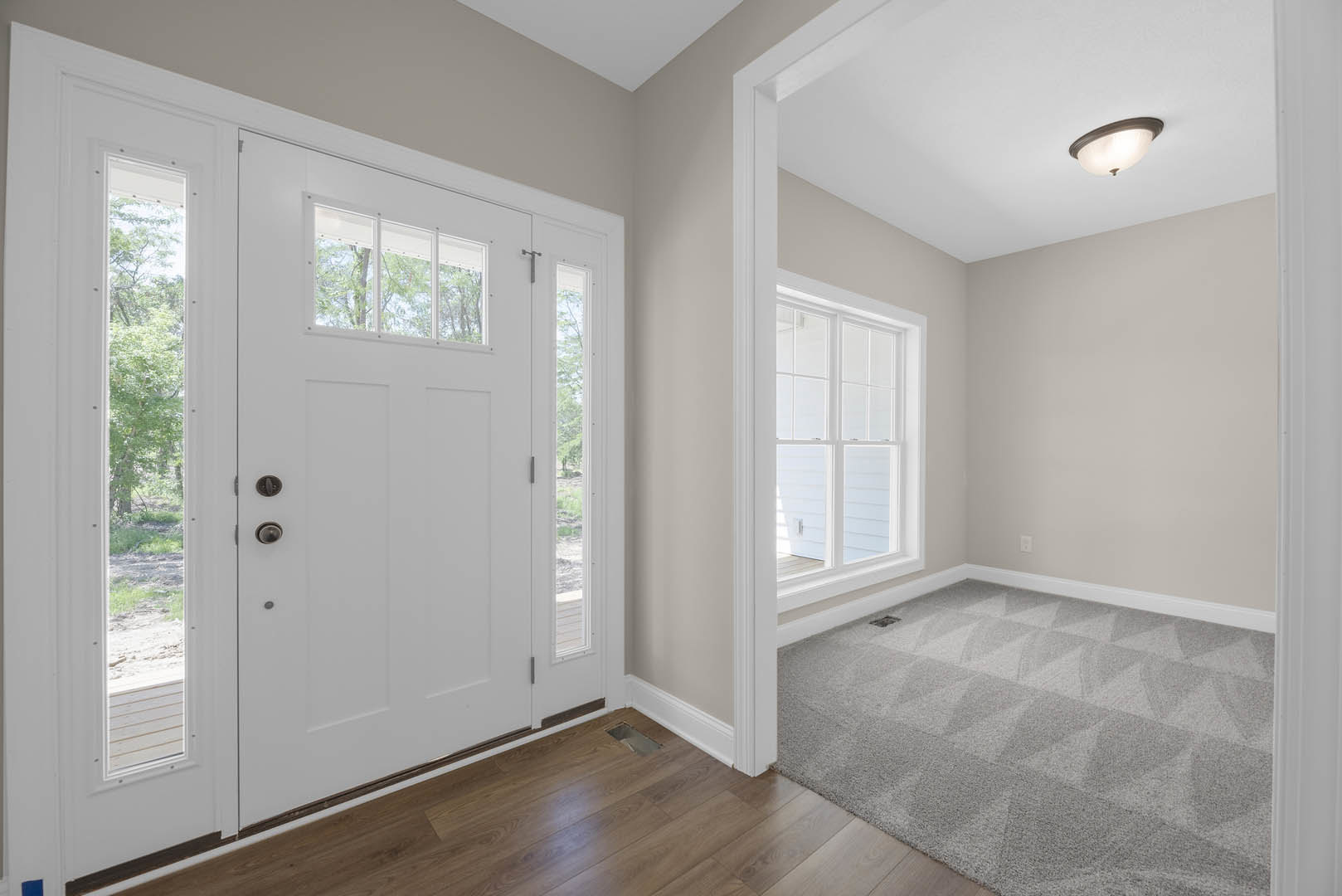 Hallway with grey carpet, white walls, white door with glass panels, recessed ceiling light, wood flooring section with cutout