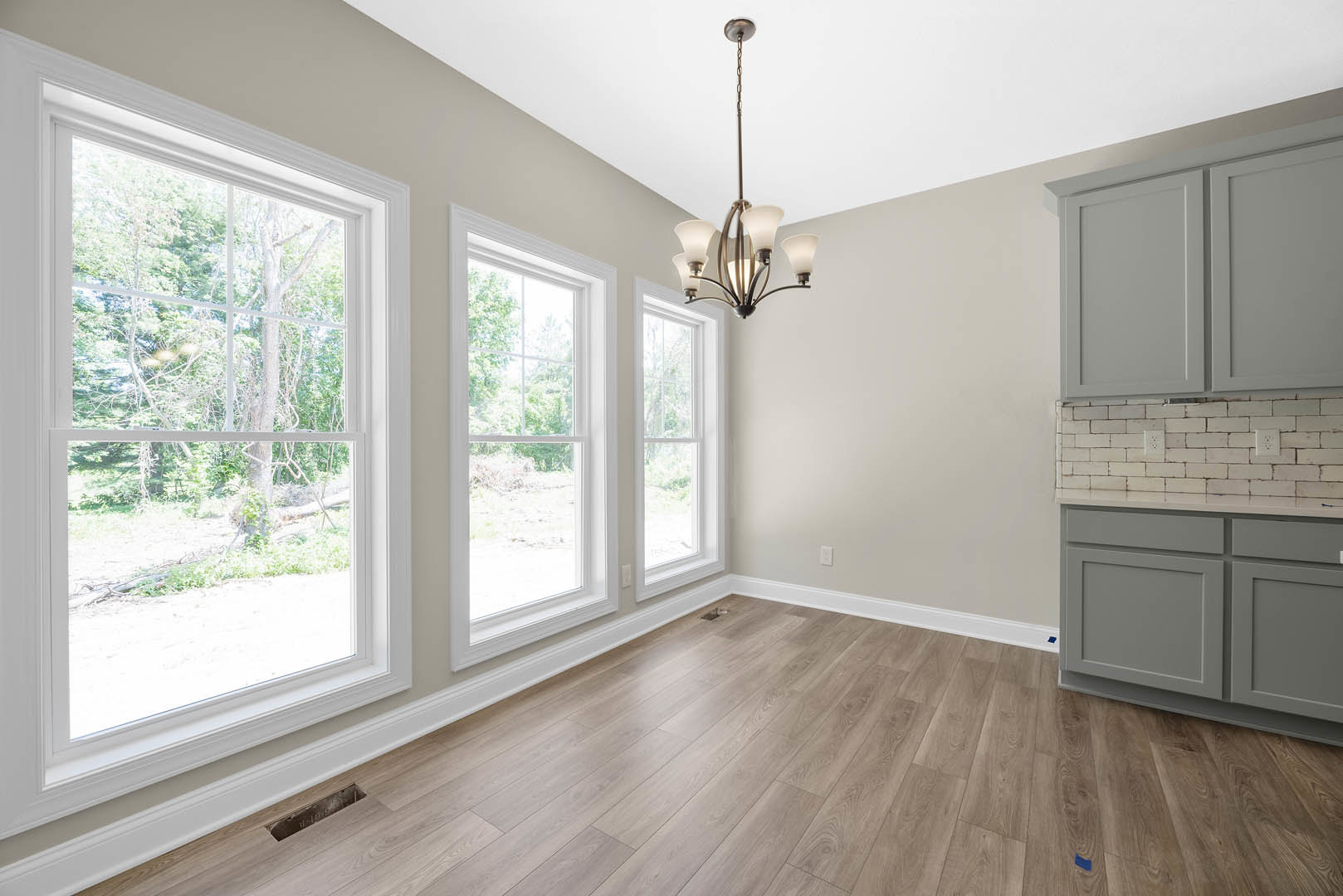 Spacious room featuring wood flooring, large windows with views of trees, elegant chandelier, white door, and modern lamp.