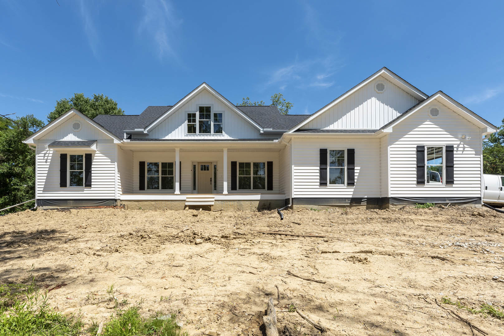Two-story house with white siding, black shuttered windows, covered front porch, and unfinished dirt yard with exposed pipe; Robert Frost Farm visible in background, tree in