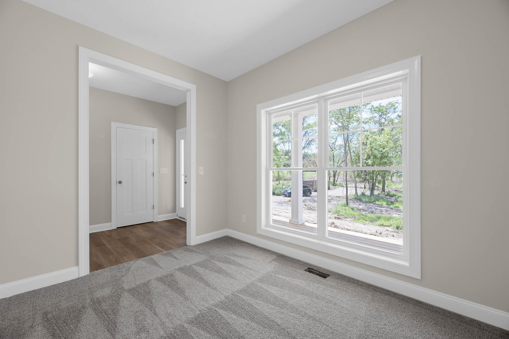Spacious room with large window overlooking trees, white door with black knob, grey carpet flooring, white walls