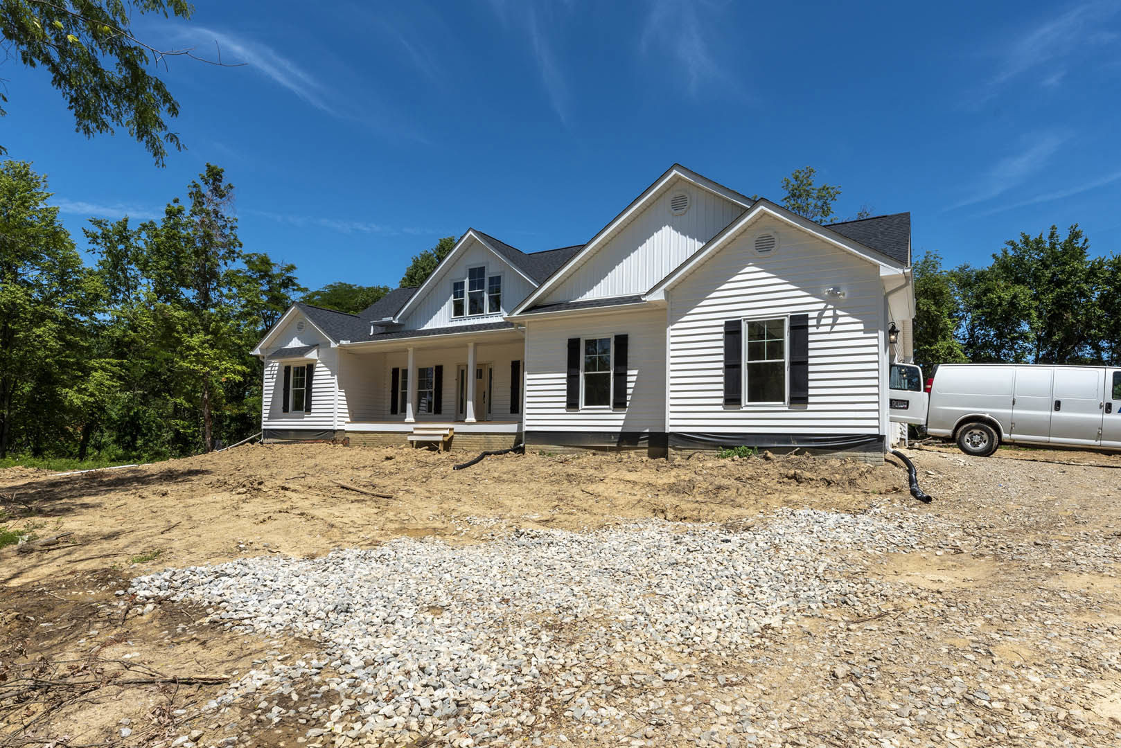Two-story home with white-framed windows and covered porch, set on a dirt hill with rocks and trees, Robert Frost Farm visible in the background under a partly cloudy sky.