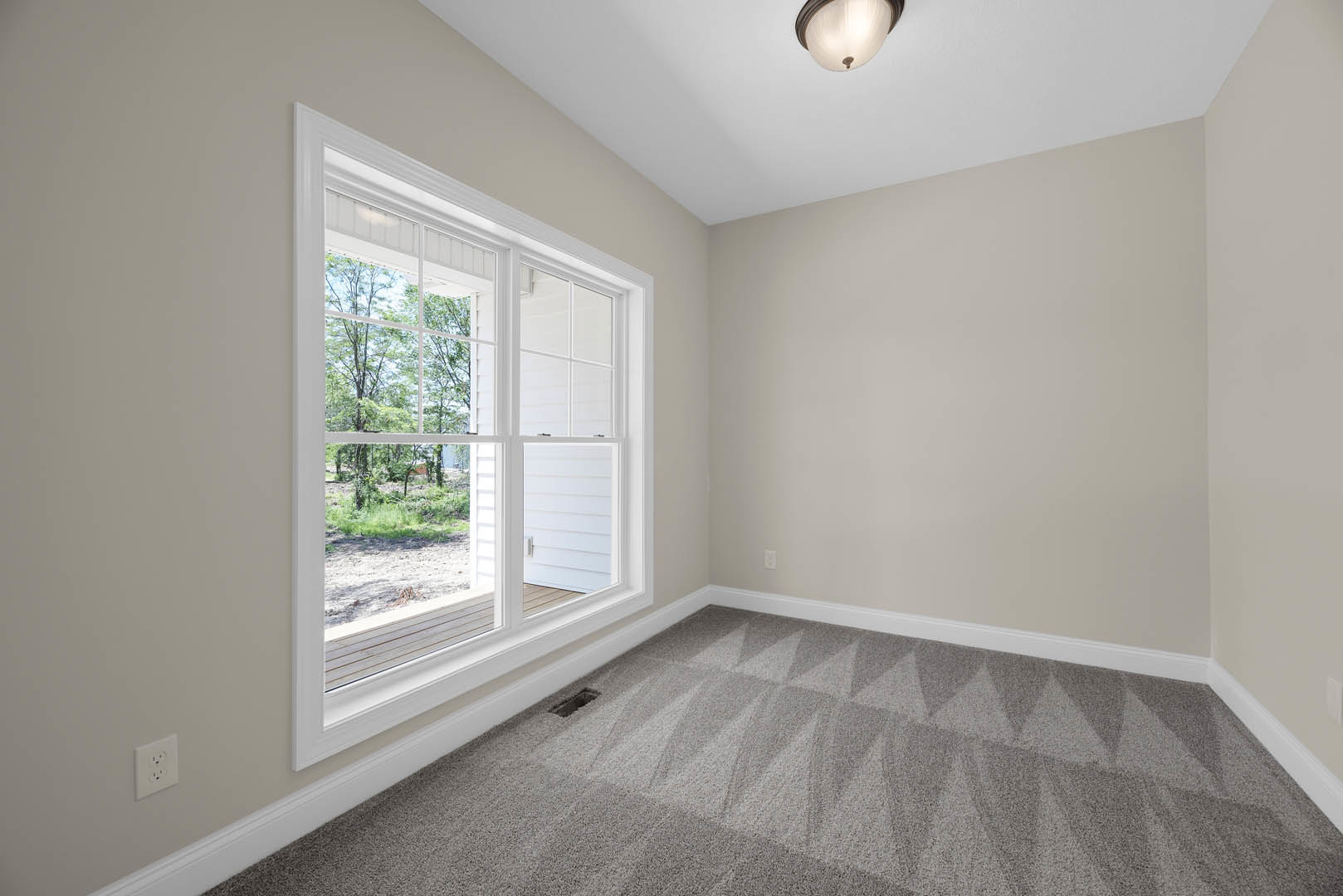 Bedroom with grey carpet, white walls, large window showing green trees, white door with matching handle, recessed ceiling light, and electrical outlet.