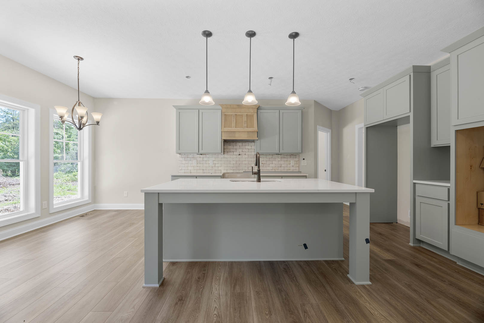 Kitchen with white island countertop, wood flooring, white cabinetry, tile backsplash, stainless steel sink and faucet, large window overlooking trees, modern chandelier, close-up