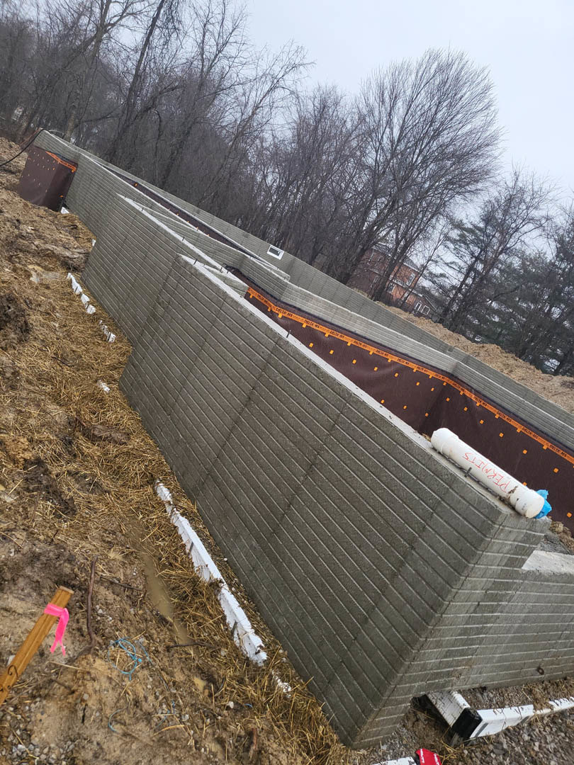Brick exterior wall atop a concrete foundation, orange construction lights and tape along the base, white cylinder with red writing placed on foundation, pink ribbon tied to a