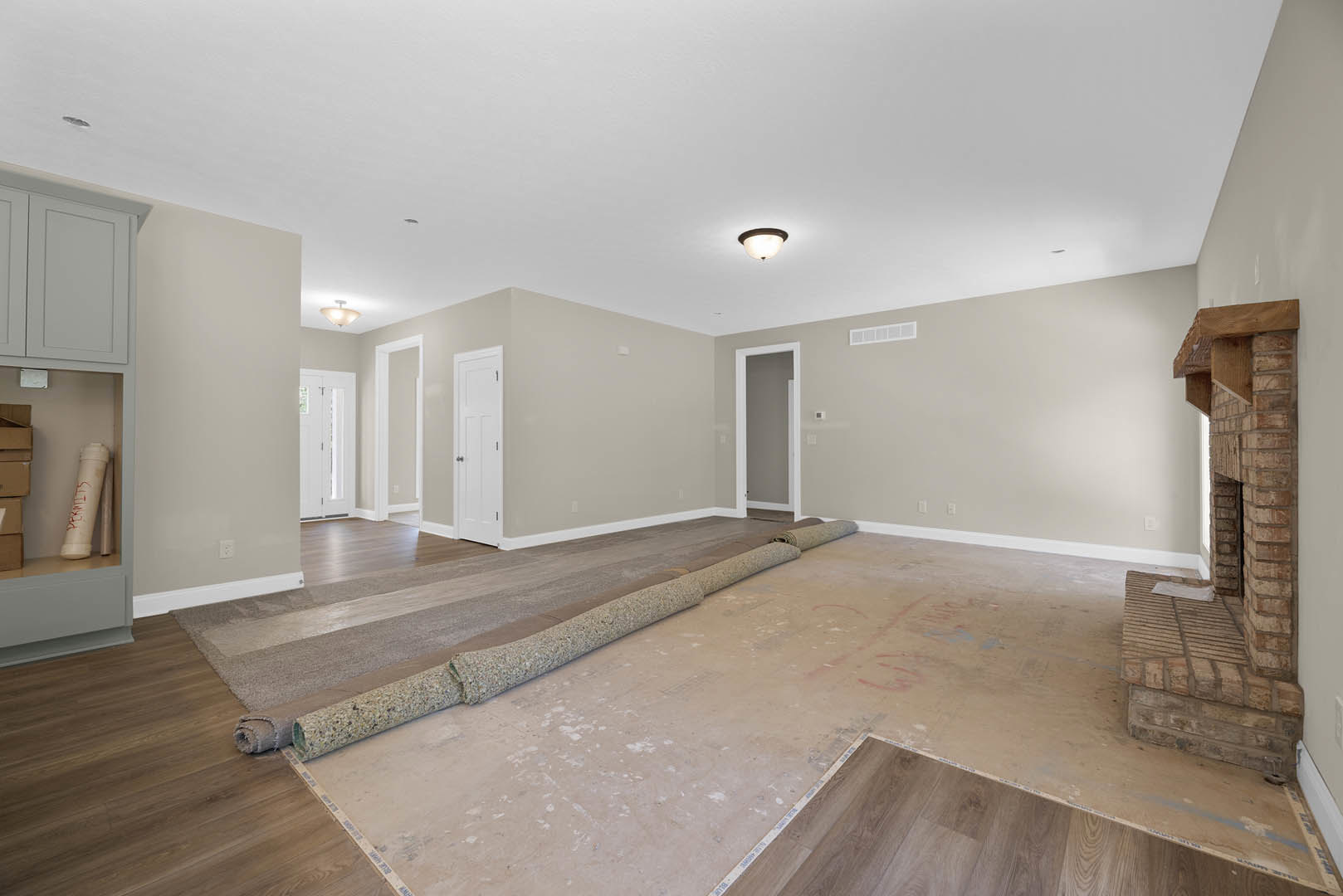 Unfinished room with rolled carpet on laminate floor, white door with silver knobs, ceiling light fixture, and exposed white pipe with red writing along plaster wall