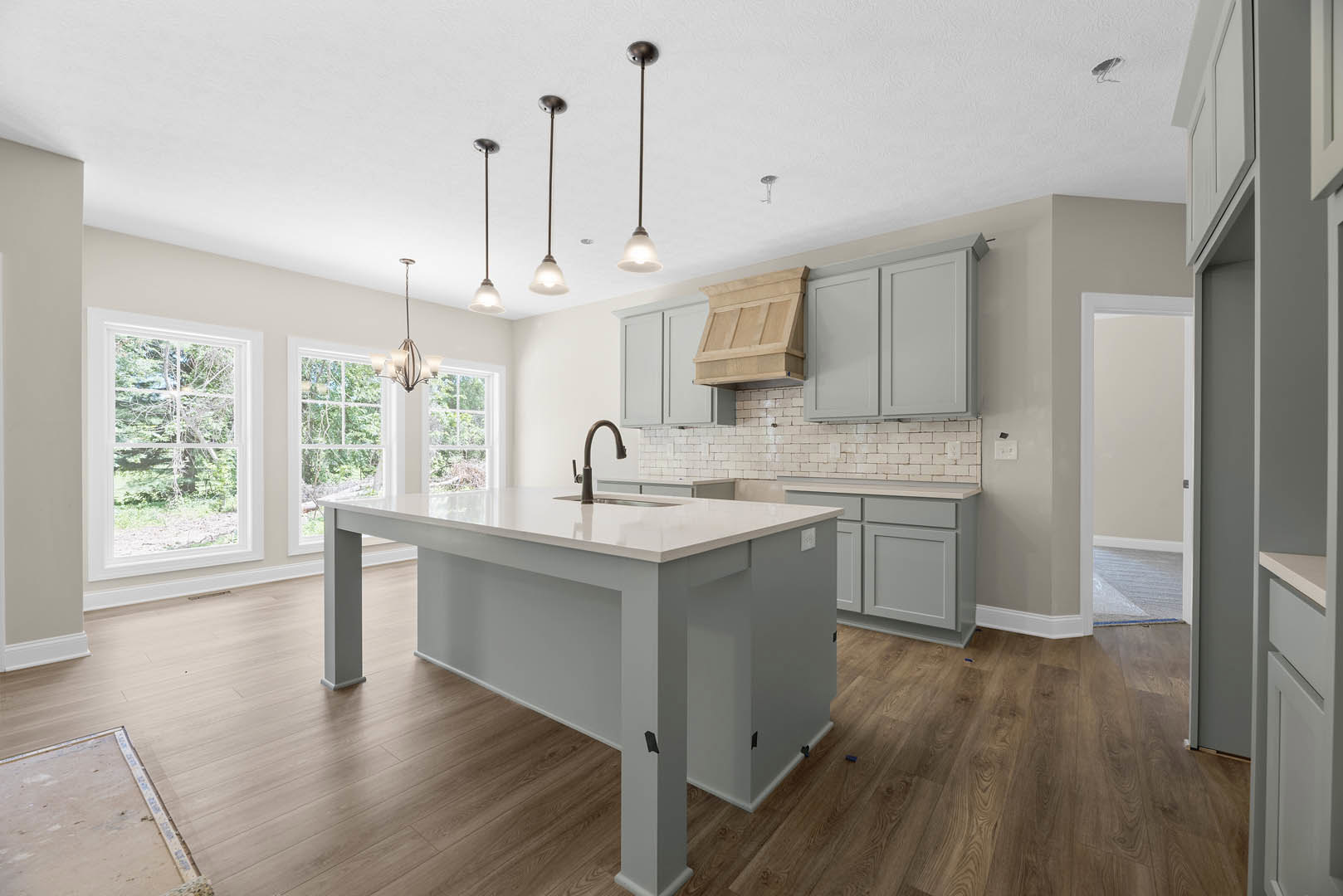 White kitchen island with black faucet, wooden vent hood, white ceiling, tile flooring, white cabinetry, window overlooking trees.