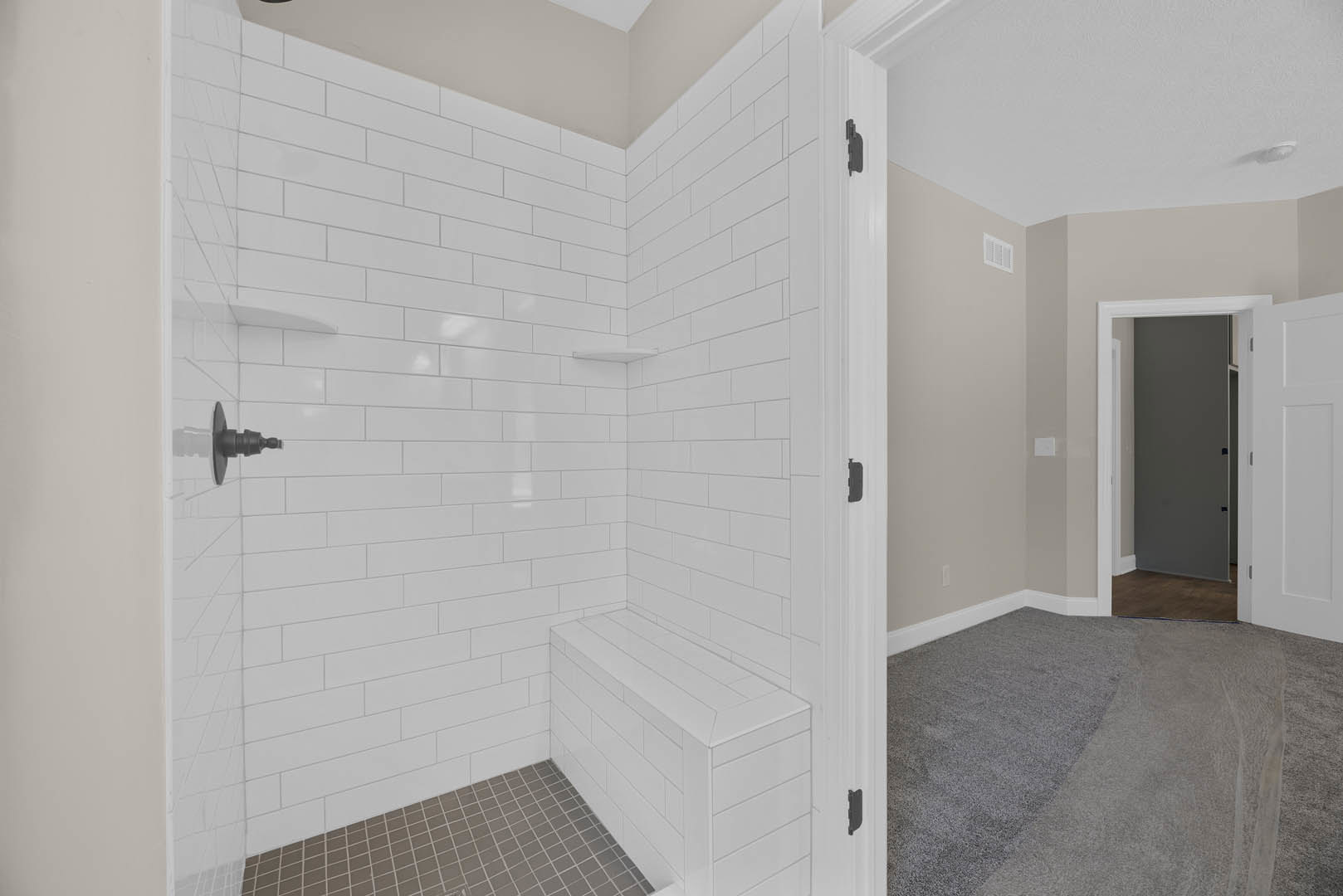 White tile shower with built-in bench and glass door, adjacent to a white door with black handle, grey carpeted floor visible in nearby room