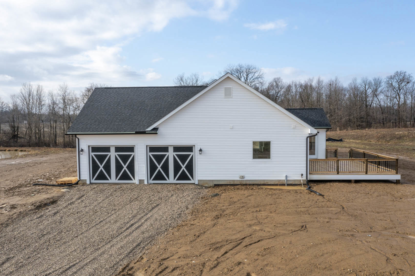 White siding house with attached garage, gravel driveway, wooden deck railing, black-framed windows, and surrounding trees under a cloudy sky