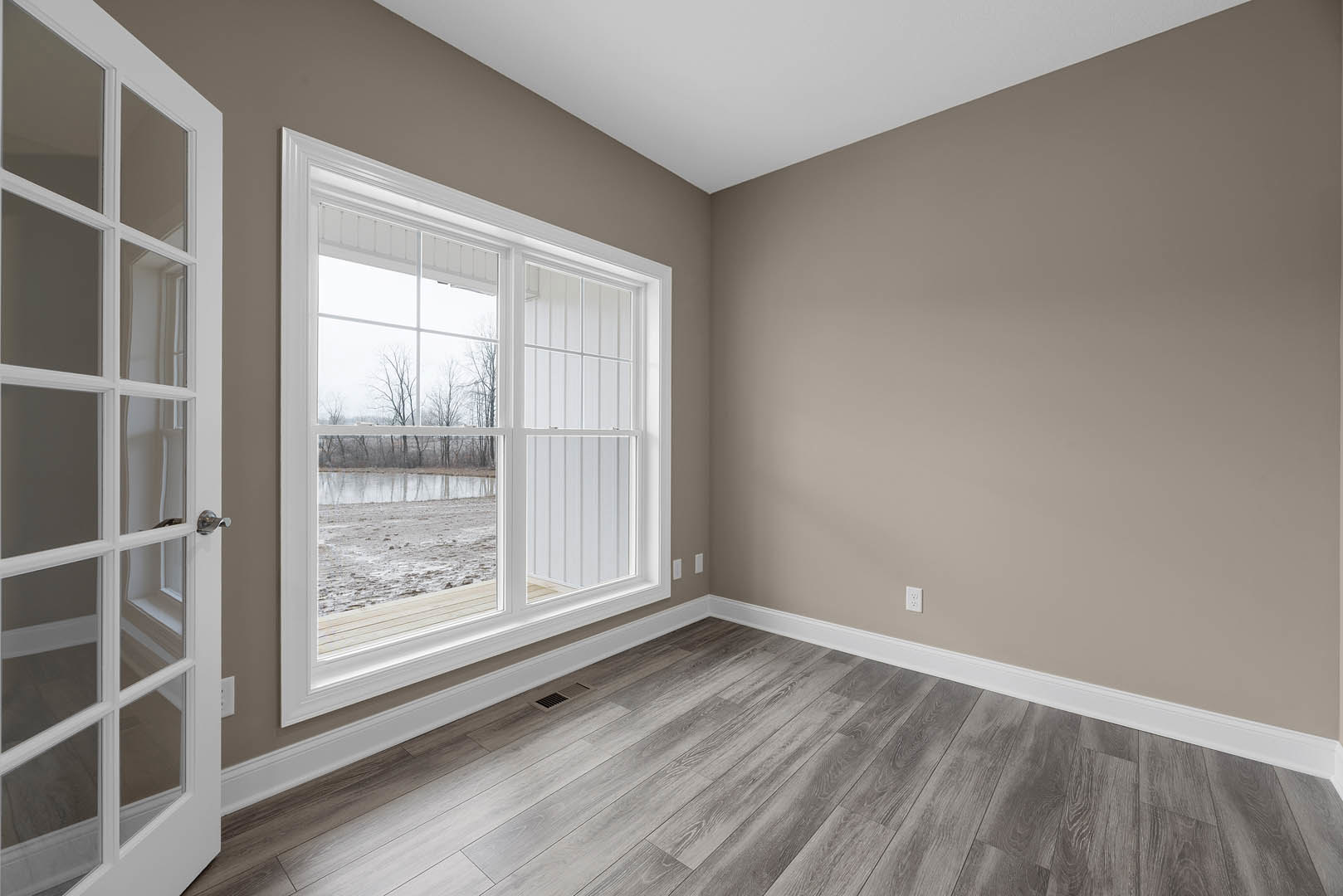 Sunlit room with white plaster walls, large window overlooking muddy field and distant trees, wood laminate flooring with floor vent near corner, partial view of white door