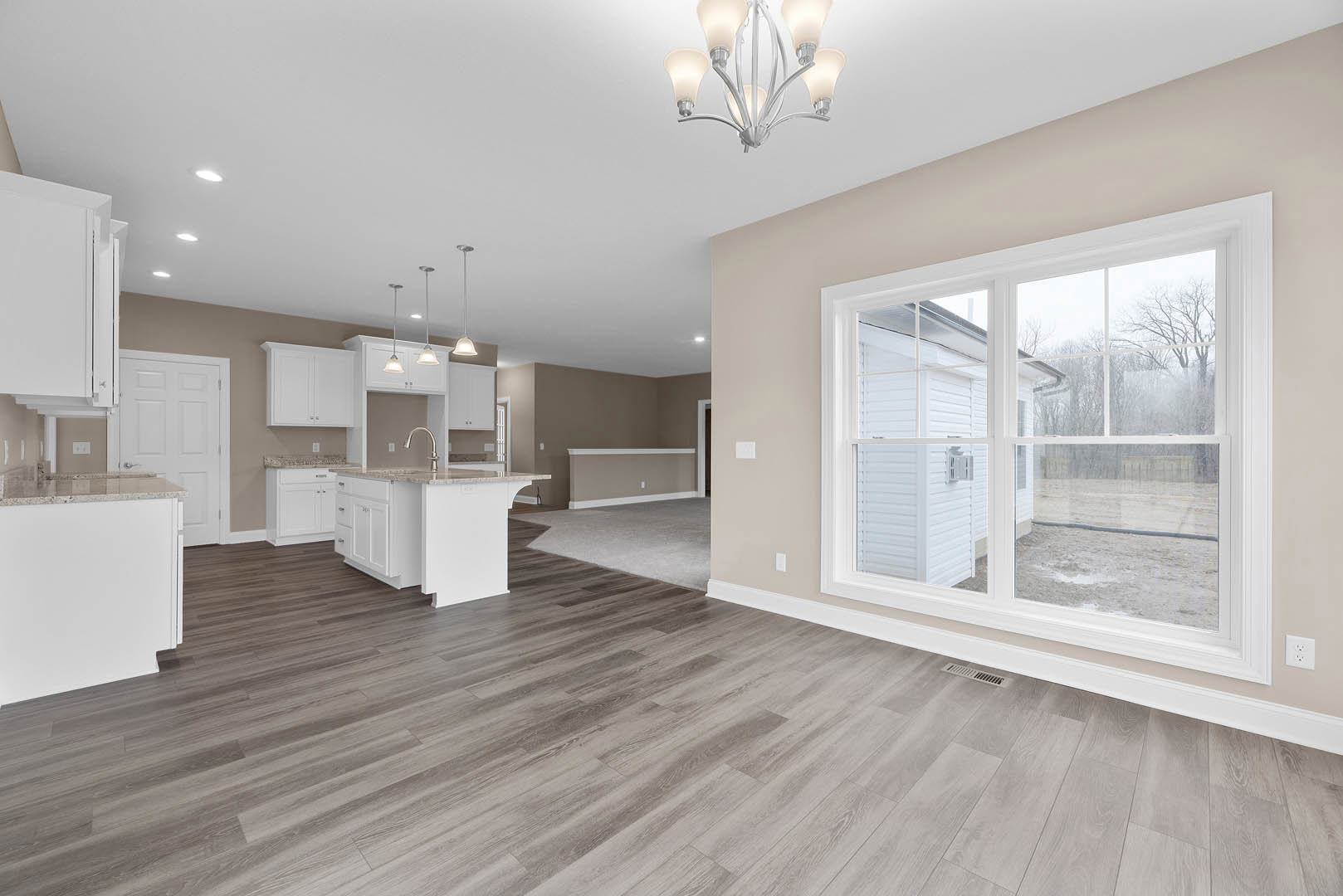 Spacious open floor plan featuring a white kitchen island with marble countertop, hardwood flooring, white cabinetry, dining area, chandelier, and large window.