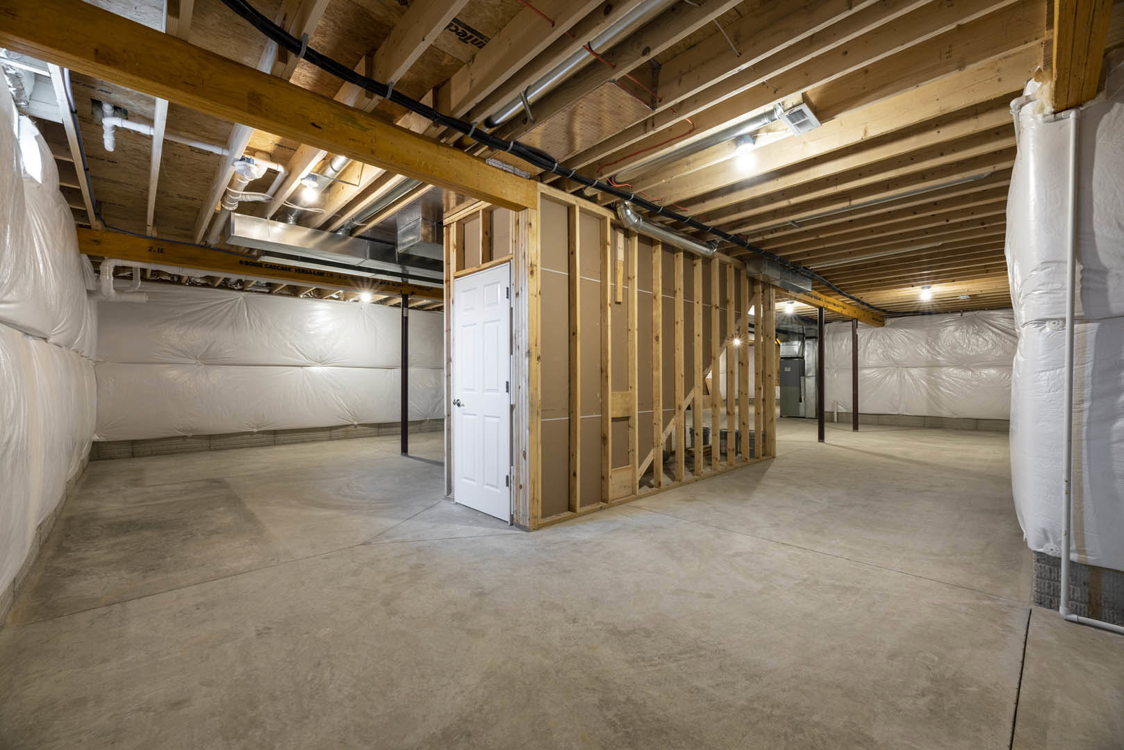 Unfinished room with exposed wood ceiling beams, visible pipes and wires, white door with metal doorknob, white plastic sheet draped over bed frame, and white plastic covering