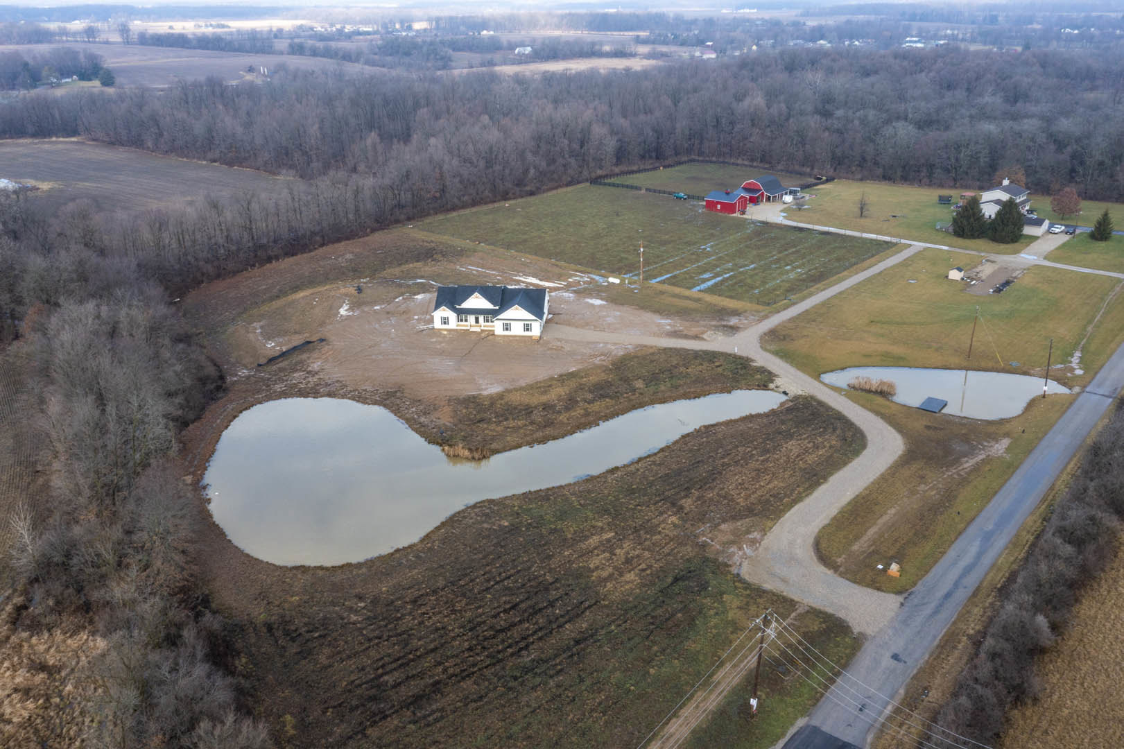 Modern farmhouse with blue roof and red barn, surrounded by grassy field, pond at center, aerial view with trees and sky in background